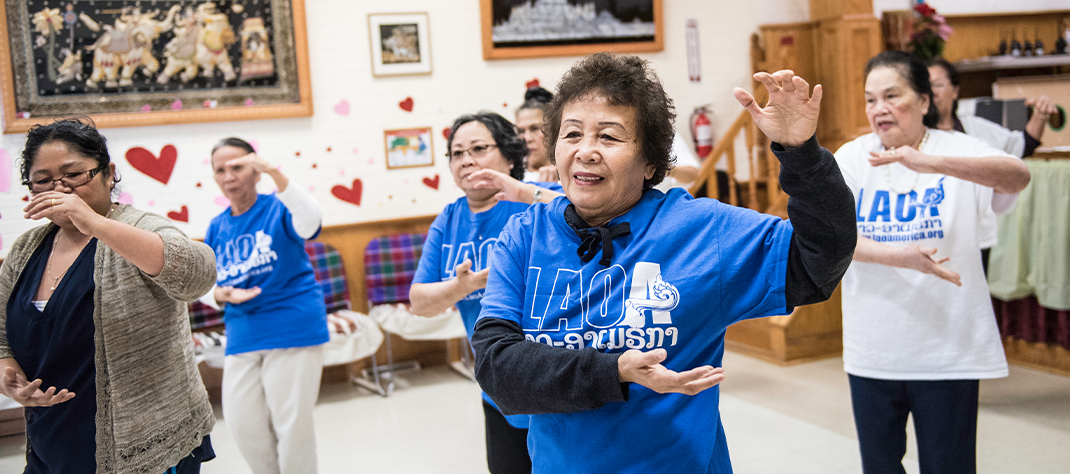 Seven older adult women doing movements with their left hand raised and their right hand across their body with the palm up. Three wear blue shirts with the letters LAOA on them for the Lao Advancement Organization of America.