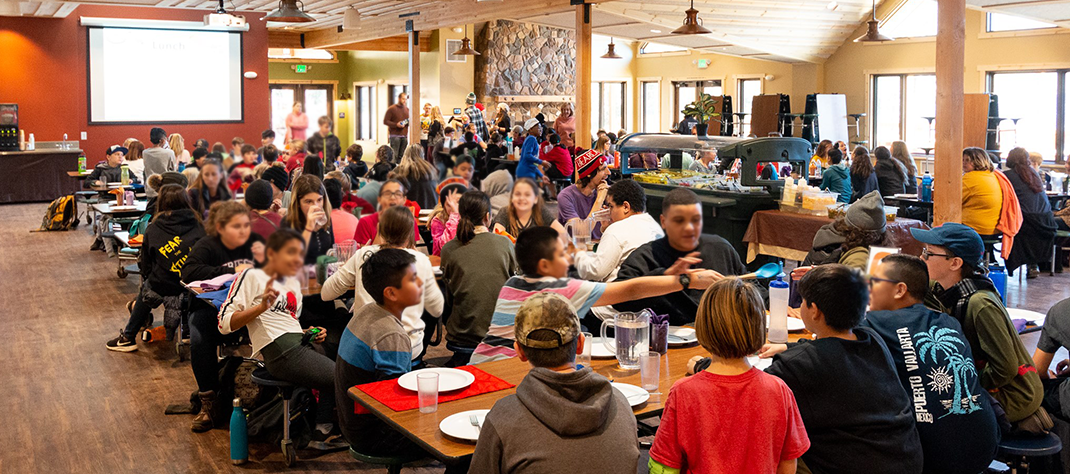 A large dining area with a wood floor and many children seated at cafeteria style tables.