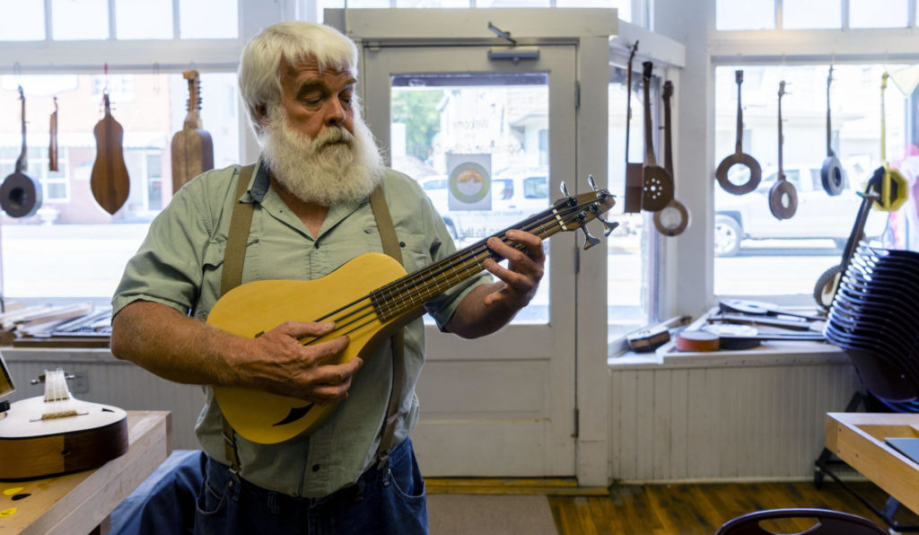 A man with white hair and beard wearing a green shirt with the sleeves rolled strums a guitar inside of a room with large windows and sunlight shining through them. The windows have eleven other stringed instruments hanging on display.