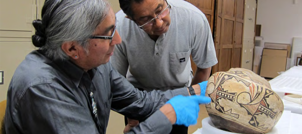 Two men look closely at an earthenware vase. The man in the forefront is holding the vase while wearing light blue gloves and both men wear glasses. The vase has geometric designs in black and reddish orange ink around the middle and it rests on a foam plate. The man in the forefront is SAR Jim Enote of the Zuni Pueblo tribe.