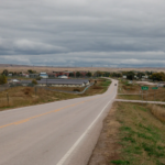 A view of the horizon and a long stretch of road into Kyle, South Dakota. There are two signs on the right of the road, a green marker with white writing that reads "Kyle" and a white sign that reads "Speed Limit 40".
