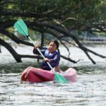 A girl in a red canoe paddling withon a river while passing under a tree.